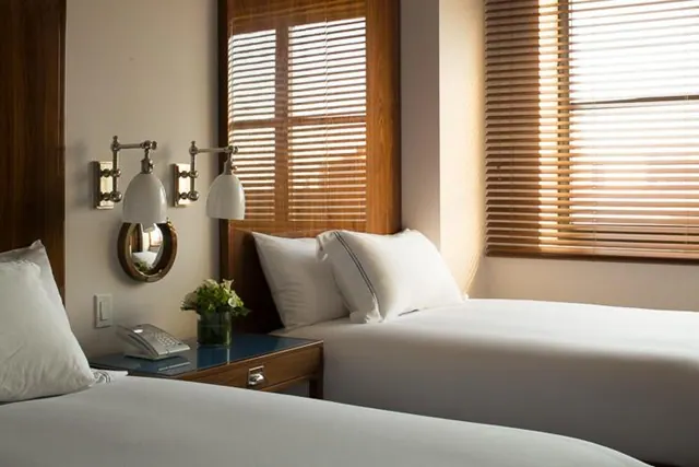 Two beds with white bedding sit beside a wooden nightstand holding a telephone and a vase of flowers, with sunlight coming through wooden window blinds.