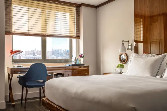 A bedroom with a made king bed, a wooden desk with a red lamp and blue chair, and a window with partly lowered bamboo blinds showing city buildings.