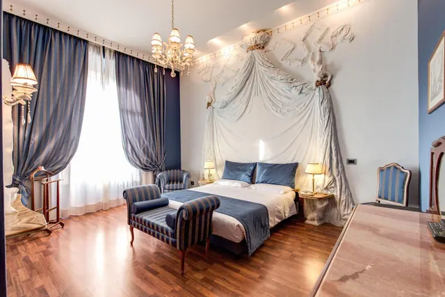 A bedroom with a double bed under a sculpted drapery headboard, a striped bench at the foot of the bed, blue striped curtains, and a crystal chandelier.