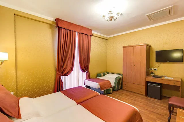 A hotel bedroom with two twin beds in the foreground, a smaller extra bed by the window, a wardrobe, a wall-mounted TV and a desk with a minibar fridge.