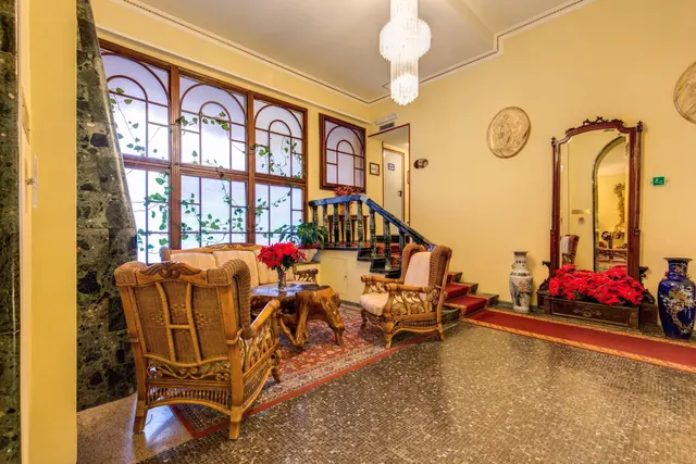 A lobby with wicker armchairs and a sofa beside tall arched windows, a staircase, a large mirror, and red poinsettias on the floor.