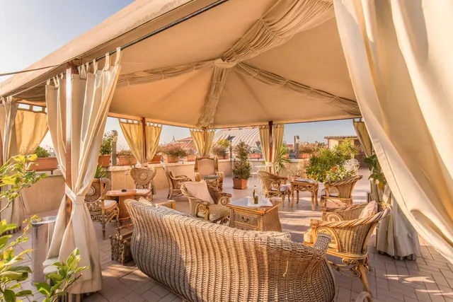 A covered rooftop terrace with wicker sofas and armchairs, potted plants and cream canvas curtains, with rooftops visible beyond.