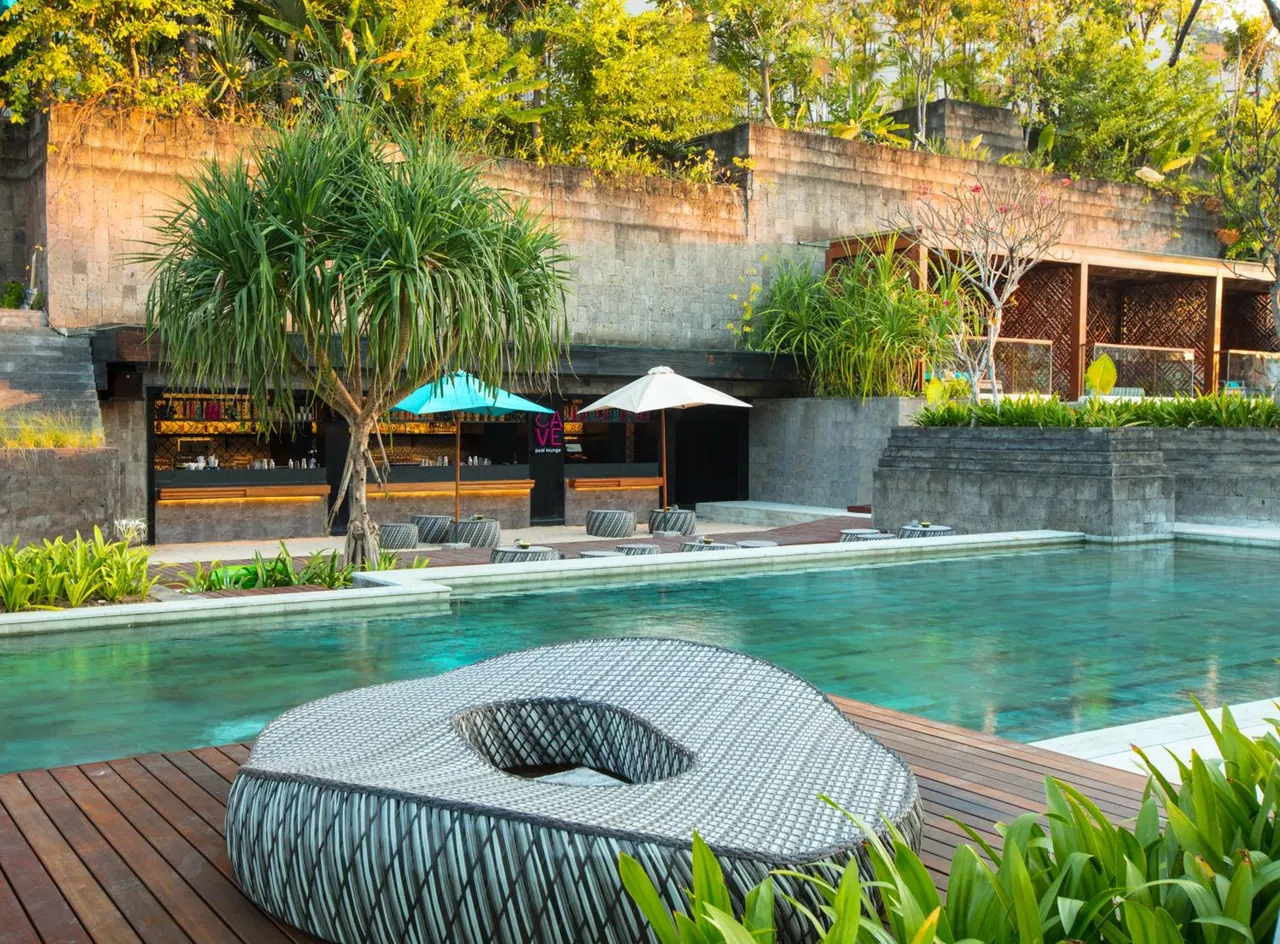 A large swimming pool with a timber deck and a woven daybed in the foreground, with a bar, umbrellas and loungers behind it.