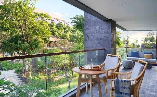 A private balcony with two rattan armchairs, a round wooden table holding a tray and bottle, and a cushioned daybed, overlooking tropical gardens and a building.
