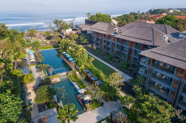 Aerial view of a resort with a pool, sun loungers and umbrellas, multi-storey buildings with balconies, and the sea beyond.