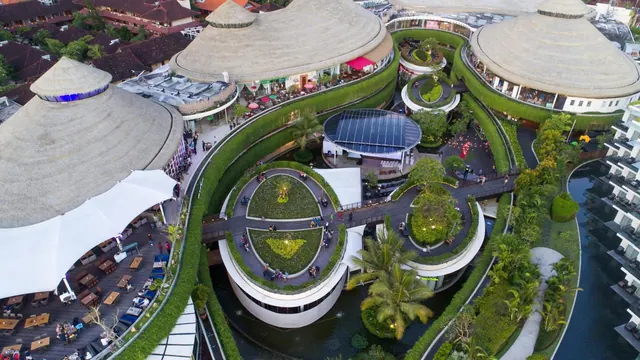 Aerial view of a complex of thatched-roof buildings with green walls, a central garden with walkways, a water channel, palm trees, and an outdoor dining area under a white canopy.