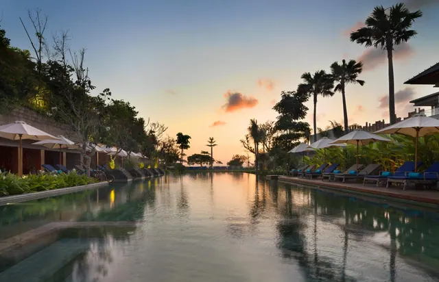 A long lap pool lined with sun loungers and parasol umbrellas under palm trees at sunset.