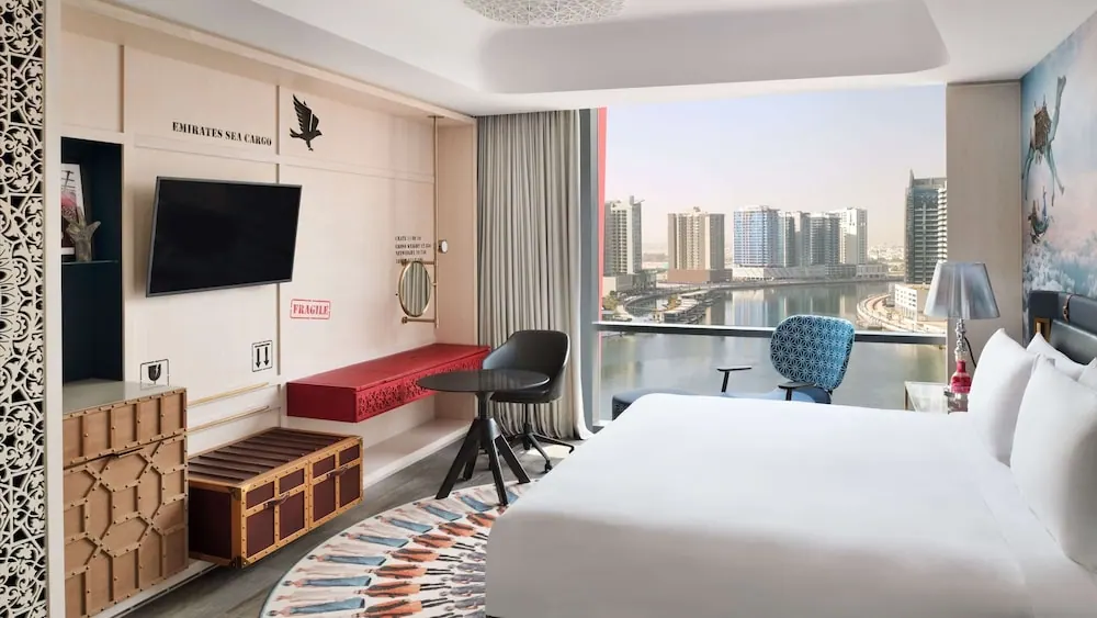 A hotel bedroom with a king bed, a round table with chairs, a wall-mounted television and a large window overlooking a canal and city towers.