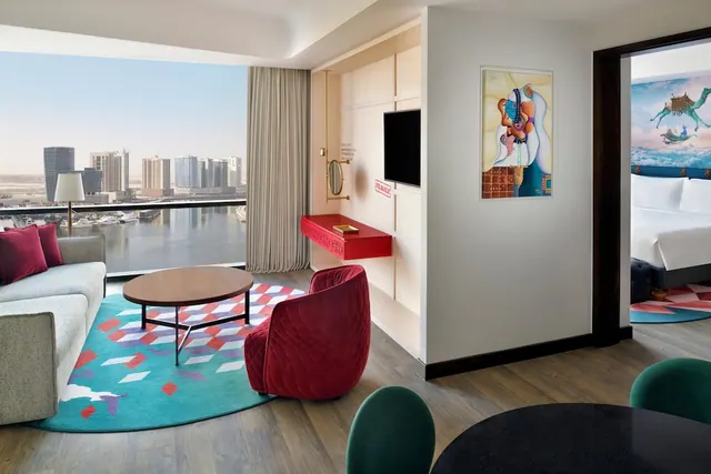 A living area with a grey sofa, round wooden coffee table, red armchair and a view of city towers and a canal through floor-to-ceiling windows, with a bed visible through an open doorway.