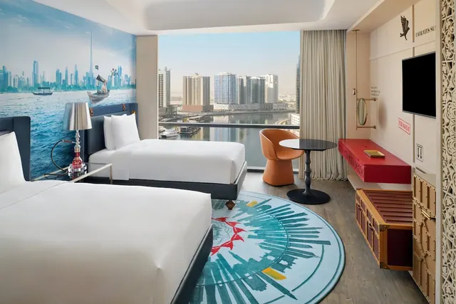 A twin-bedded hotel room with a Dubai skyline mural on the wall and a window view over a canal lined with high-rise buildings.