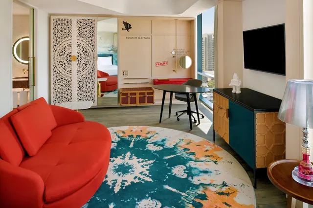 A living area with a red curved sofa, a round patterned rug, a desk, a wall-mounted television and a window showing high-rise buildings, with a bedroom visible through a mirrored wardrobe.