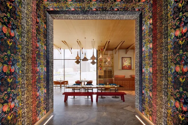 A dining room with long tables, a red bench and pendant lamps, seen through an ornately patterned doorway.