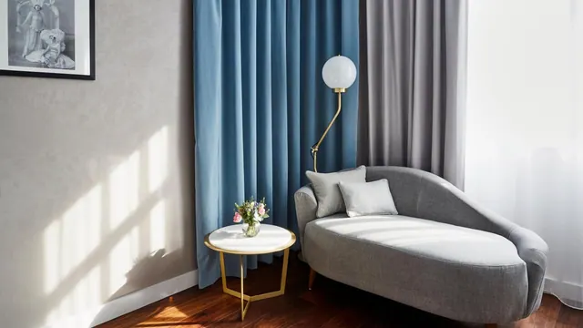 A grey chaise lounge with cushions sits beside blue curtains, a gold side table with flowers and a wall lamp, on a wooden floor.