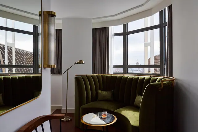 A curved green velvet banquette sits in a bay-window corner of a room beside a brass floor lamp, with rooftops and distant buildings visible through the windows.