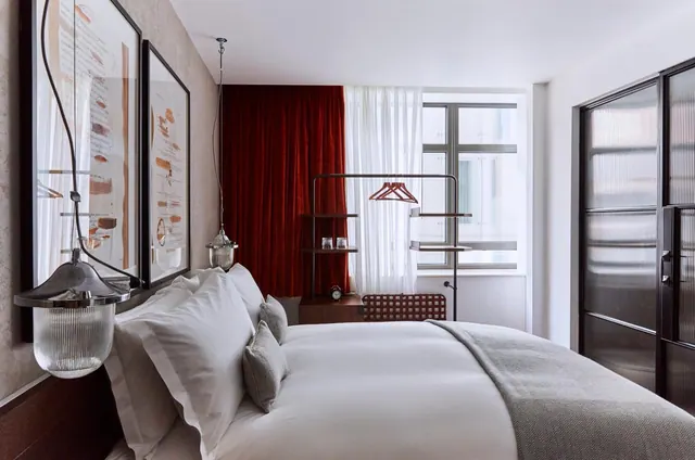 A bedroom with a made bed with white linen and a grey throw, red velvet curtains, and a window facing a neighbouring building.