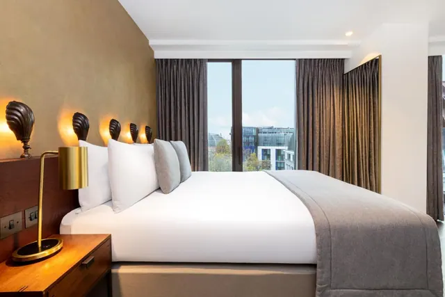 A made king bed with white linen and a grey throw sits beside a large window looking out onto buildings and trees.