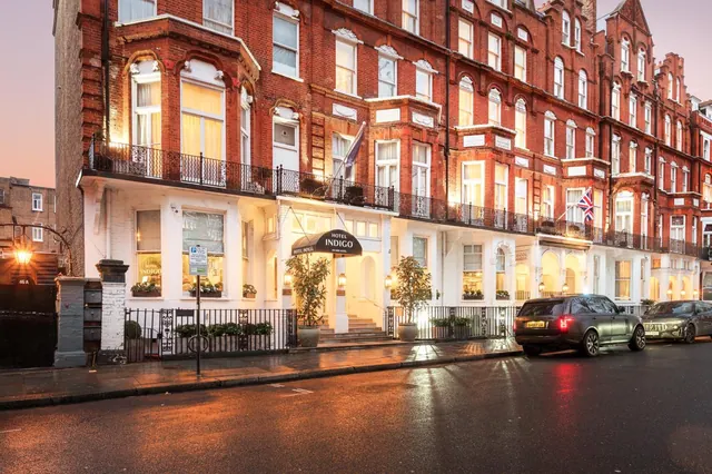 Red brick Victorian facade of Hotel Indigo at dusk, with lit ground-floor windows, a dark entrance canopy and parked cars on the wet street.