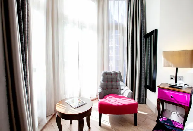 A seating corner with a grey tufted armchair, a pink ottoman, a round wooden side table and a pink bedside table beside sheer-curtained windows.