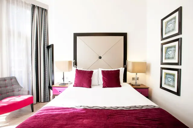 A bedroom with a double bed dressed in white linen and magenta cushions, flanked by pink bedside tables and lamps, with an upholstered armchair and a pink bench by the window.