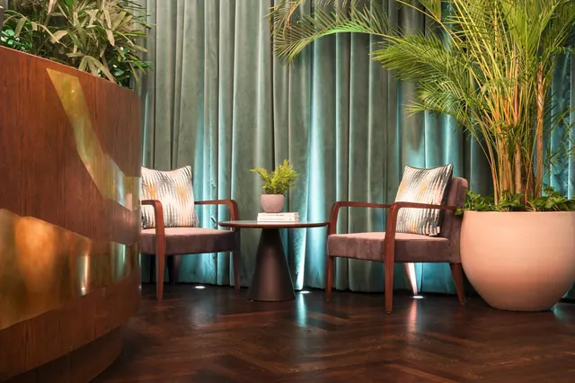 Two upholstered armchairs flank a small round side table holding books and a small plant, with teal velvet curtains and a large potted palm beside them on a herringbone wood floor.
