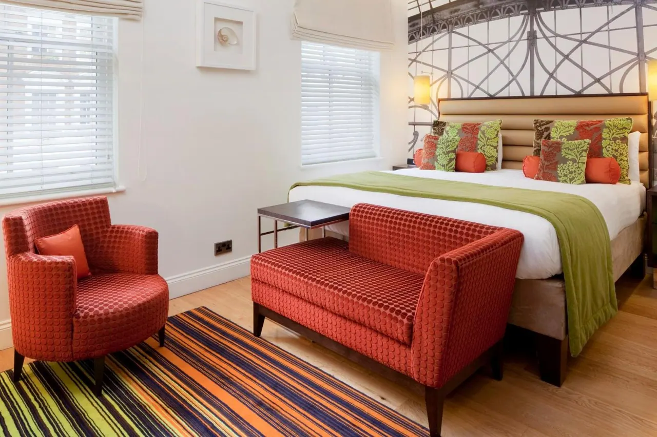A bedroom with a made double bed, a red upholstered bench at its foot, a red armchair by the window and a striped rug on the wooden floor.