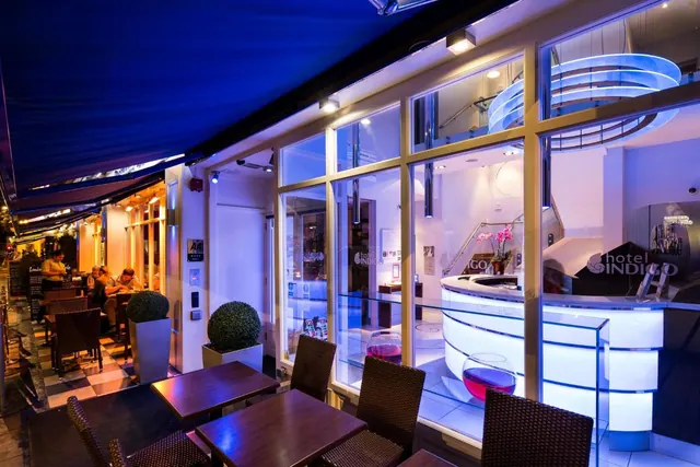 Evening view of the Hotel Indigo entrance, showing outdoor tables under a blue awning and the reception desk seen through large glass windows.