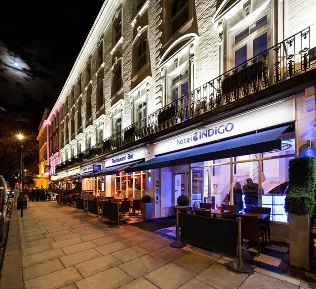The lit front of a Hotel Indigo building at night, with tables and chairs set out on the pavement beside a Restaurant Bar sign.