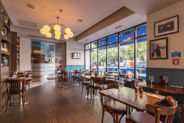 A dining room with wooden bentwood chairs and tables, a globe chandelier, and large windows looking onto a street with parked cars and trees.