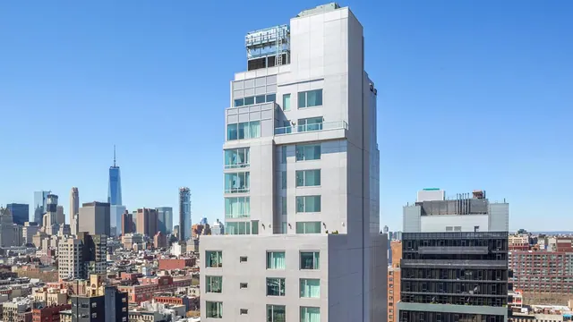 Exterior of a tall white hotel tower under a clear blue sky, with a city skyline including One World Trade Center behind it.