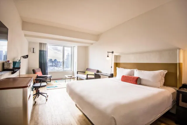 A king bed with a white duvet and red bolster faces a desk, armchair and a window looking out onto city buildings.