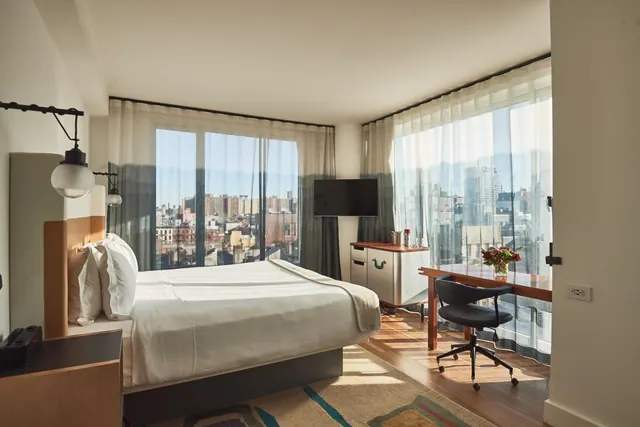 A bedroom with a made bed, a wall-mounted television, a desk with a chair, and floor-to-ceiling windows overlooking city buildings.