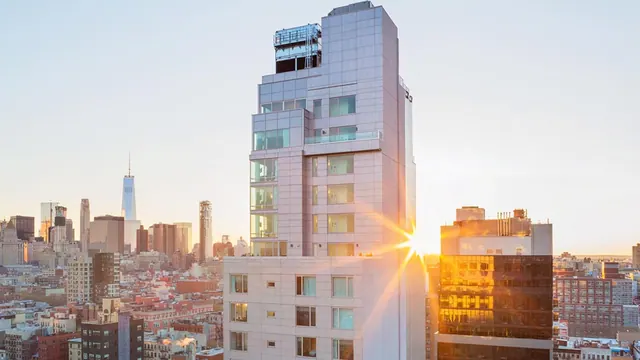 A tall light-coloured tower rises in front of a city skyline at sunset, with One World Trade Center visible in the distance.