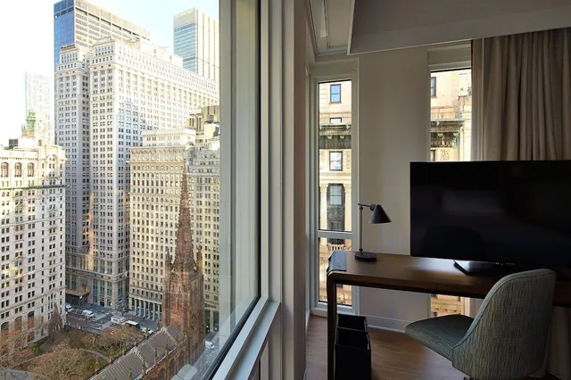 A hotel room with a wooden desk, a black lamp, a wall-mounted television and a chair beside a window overlooking high-rise buildings and the spire of Trinity Church.