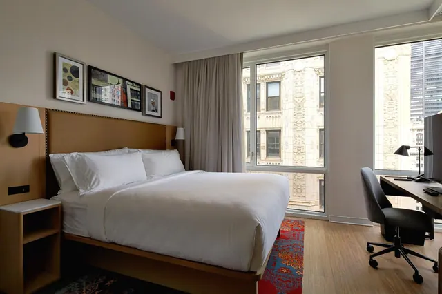 A bedroom with a king bed, a work desk and chair, and windows looking out onto an ornate building facade.