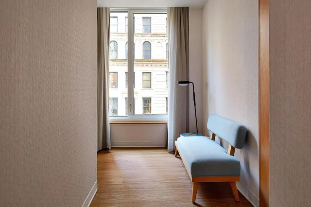 A light blue upholstered bench and a black floor lamp sit in a corner beside a window looking out onto an older multi-storey building.