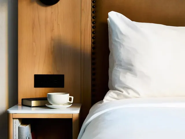 A made bed with white linens and pillows sits beside a wood-panelled nightstand holding a cup and saucer, a book, and a wall-mounted reading light.