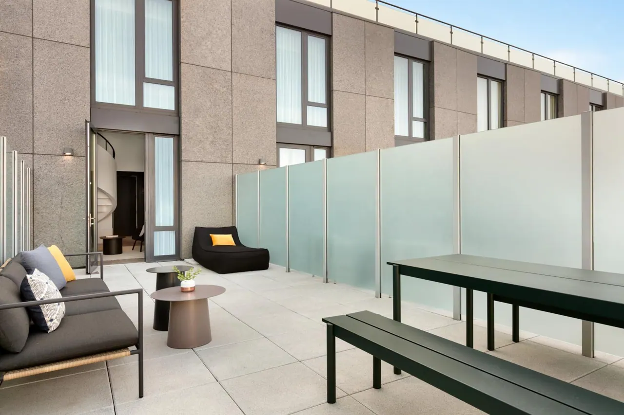 A furnished rooftop terrace with a grey sofa, low tables, a black bench and table, and frosted glass screens along the building wall.