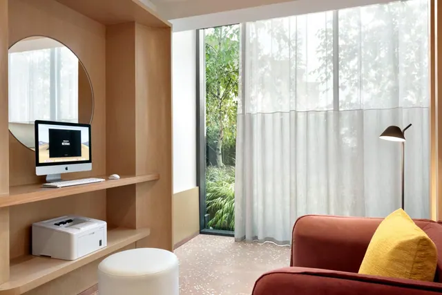 A living area with a built-in wooden desk holding an iMac and printer, a white stool, a red sofa with a yellow cushion, and sheer curtains beside a window onto green trees.