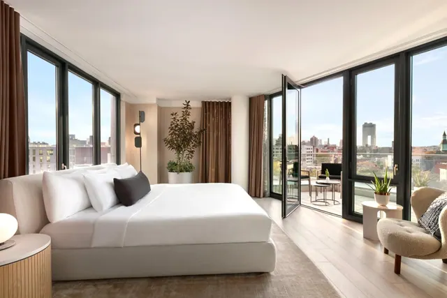 A bedroom with a made king bed, a seating chair, and floor-to-ceiling windows with open doors onto a balcony overlooking a city skyline.