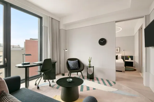 A living area with floor-to-ceiling windows, a desk and chairs, a round coffee table, and a glimpse of a bedroom through a doorway.