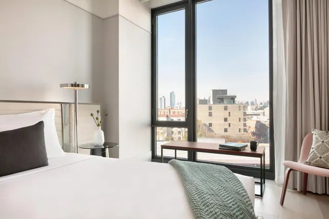 A made bed sits beside a floor-to-ceiling window looking out over city buildings and distant towers under a clear sky.