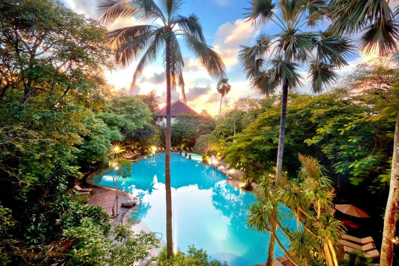A large turquoise pool is surrounded by palm trees and lush greenery at sunset, with sun loungers and umbrellas along its edge.