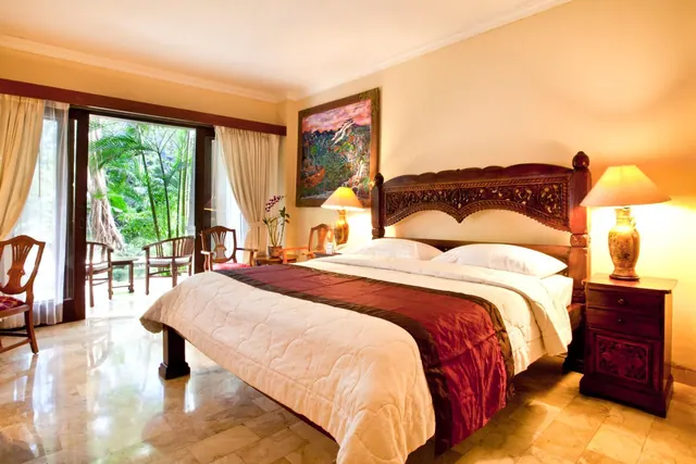 A bedroom with a carved wooden king bed, bedside lamps and armchairs, with glass doors opening onto a terrace and tropical garden greenery.