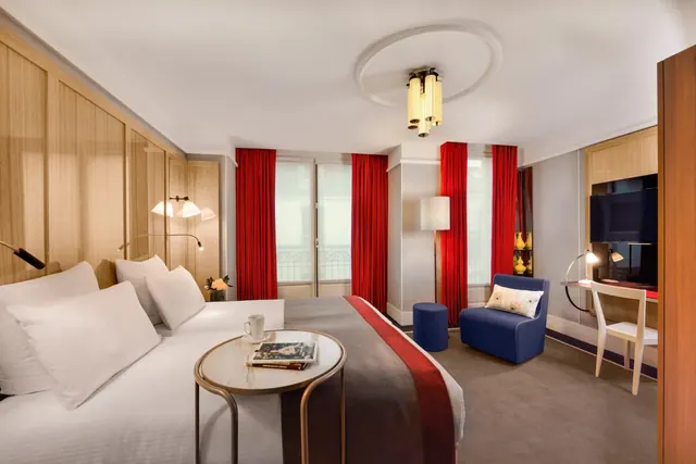 A well-appointed bedroom contains a large bed, seating area, desk, television and balcony doors framed by red curtains.