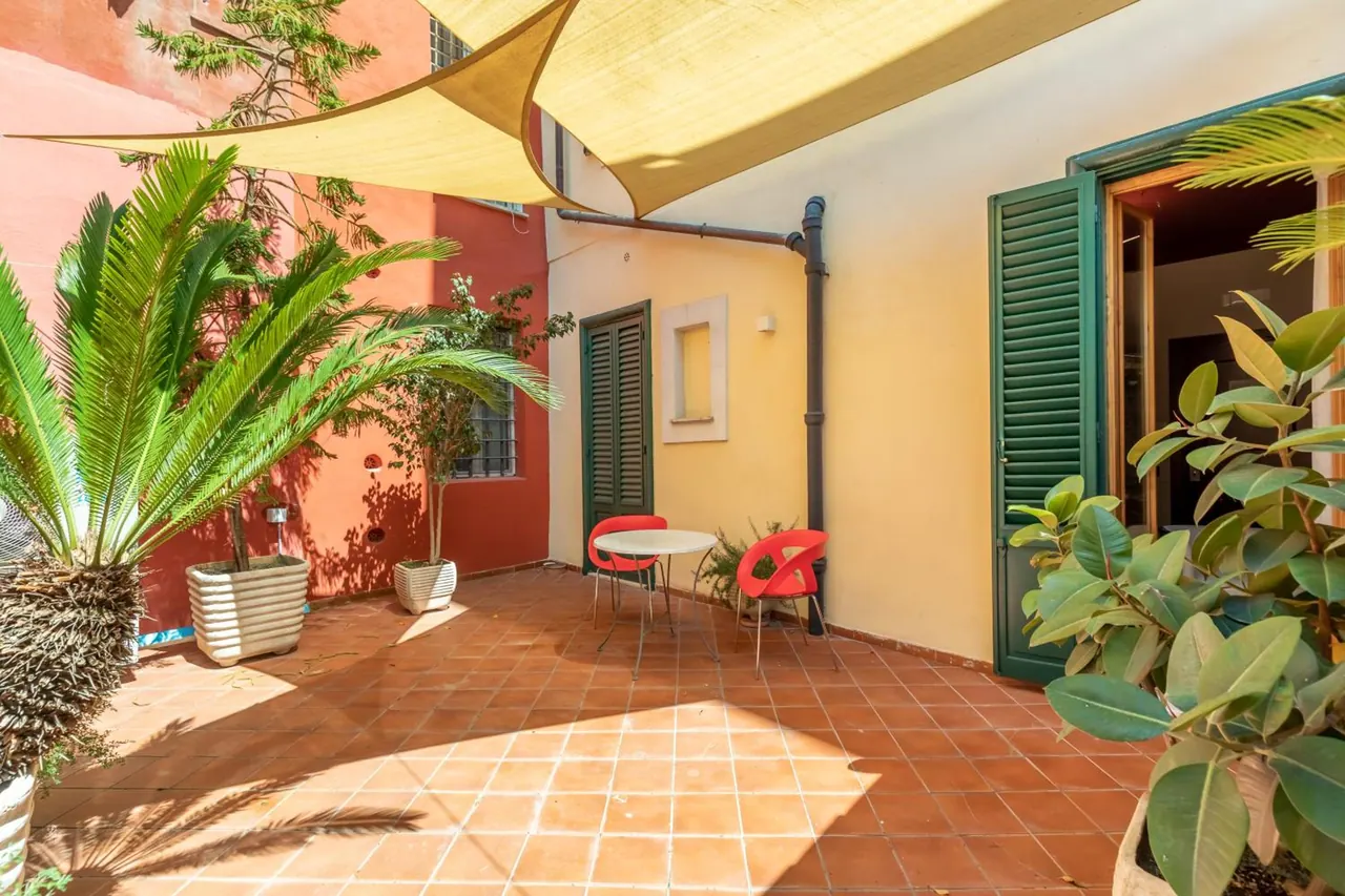 A terracotta-tiled courtyard terrace has a small round table with two red chairs, potted plants, and green-shuttered doors, under a fabric shade sail.