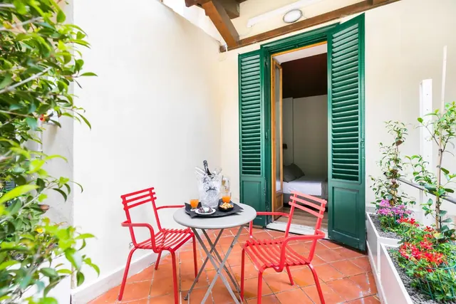 A small tiled terrace with red metal chairs, a round table holding drinks, and flower planters, with an open green-shuttered door leading into a bedroom.