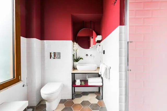 A bathroom with red upper walls and white subway tile below, a round mirror, a vessel sink on a black metal stand, a toilet, a hexagonal tiled floor, and a glass-doored shower with pink tiles.