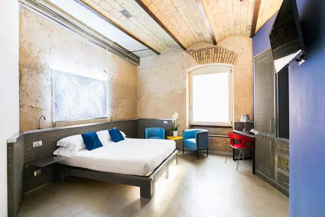A bedroom with a double bed with blue cushions, two blue armchairs, a desk with a red chair, a wall-mounted television and a vaulted brick ceiling.