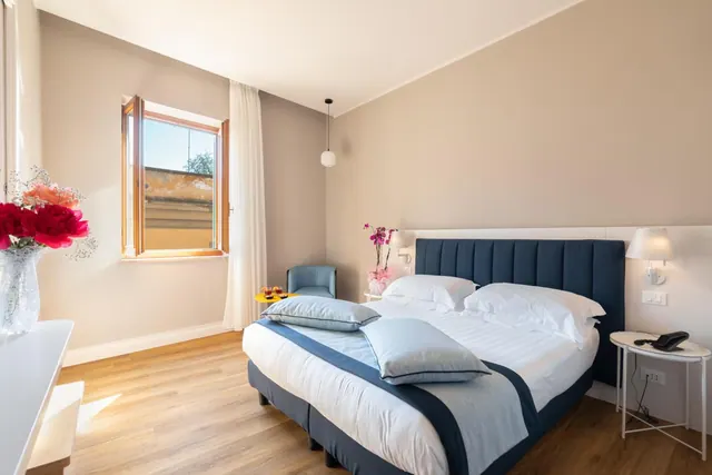 A double bed with a navy headboard and blue bedding fills a bedroom with an armchair, a window with wooden shutters, and a telephone on the bedside table.