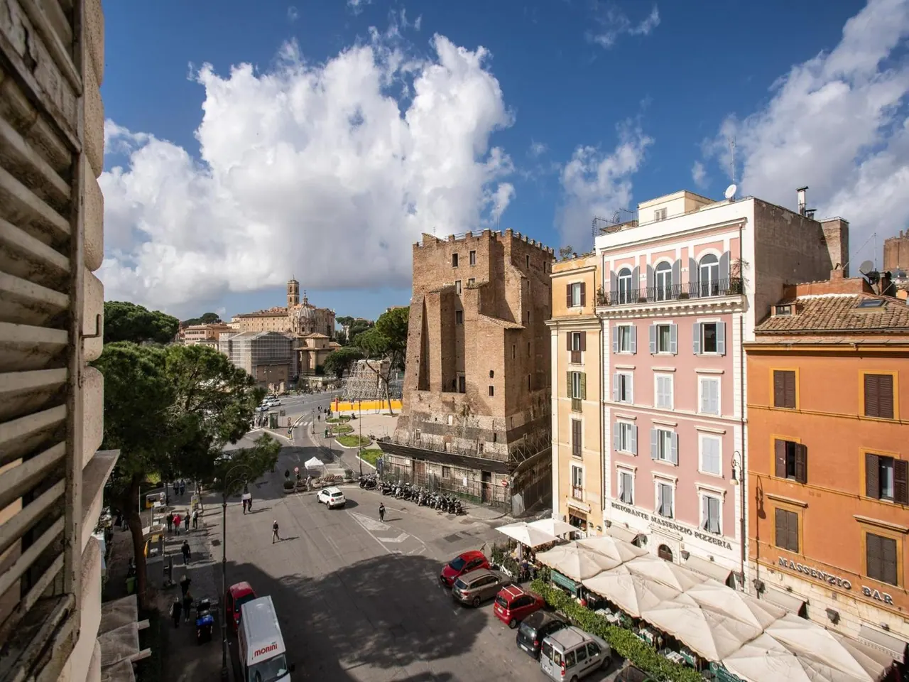View from an upper window down a Rome street toward a brick tower with scaffolding, parked cars, and a restaurant terrace with umbrellas below.